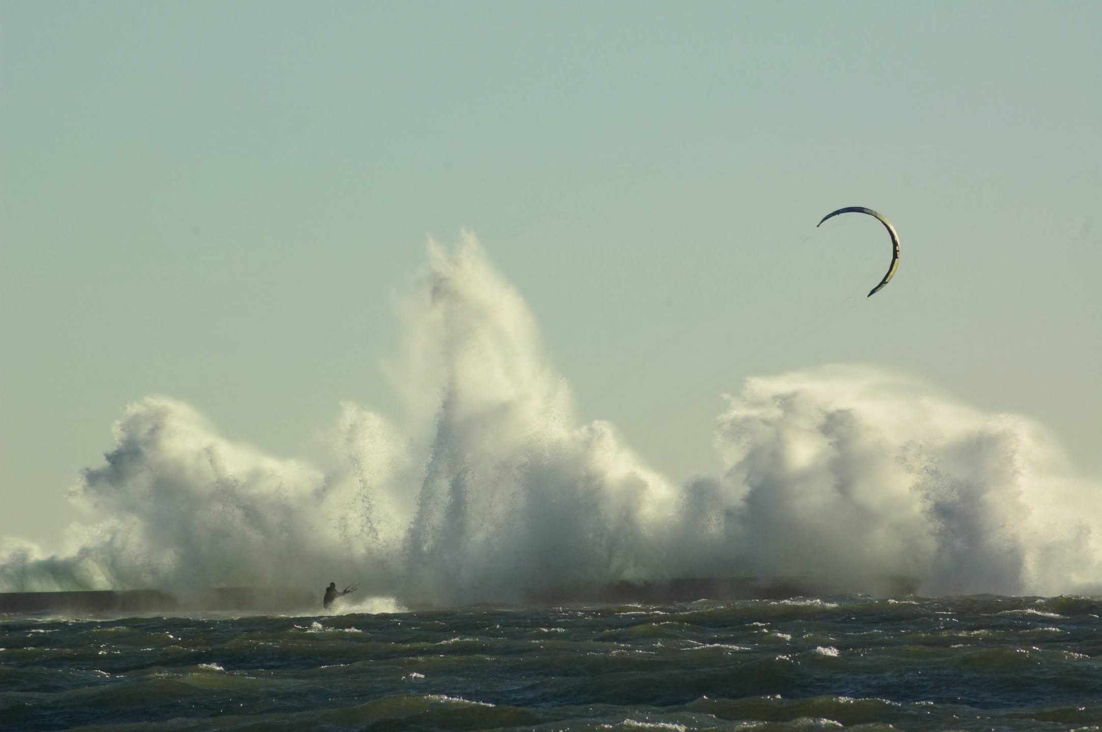 A lone kiteboarder riding past a pier as big waves explode over it