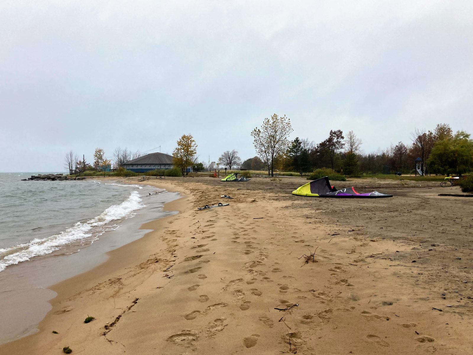 Two kites and twin-tip boards laid out on the wide sandy day-use beach at Tawas Point State Park, with the beach pavilion and autumn trees behind