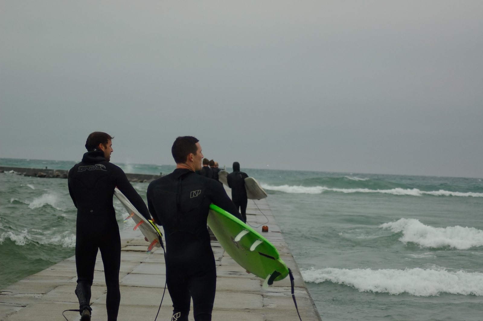 Two surfers in wetsuits carrying boards out along the Frankfort pier, waves breaking alongside